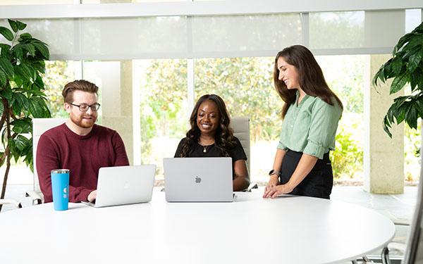 Three UWF community members gather around two laptops in a meeting room.